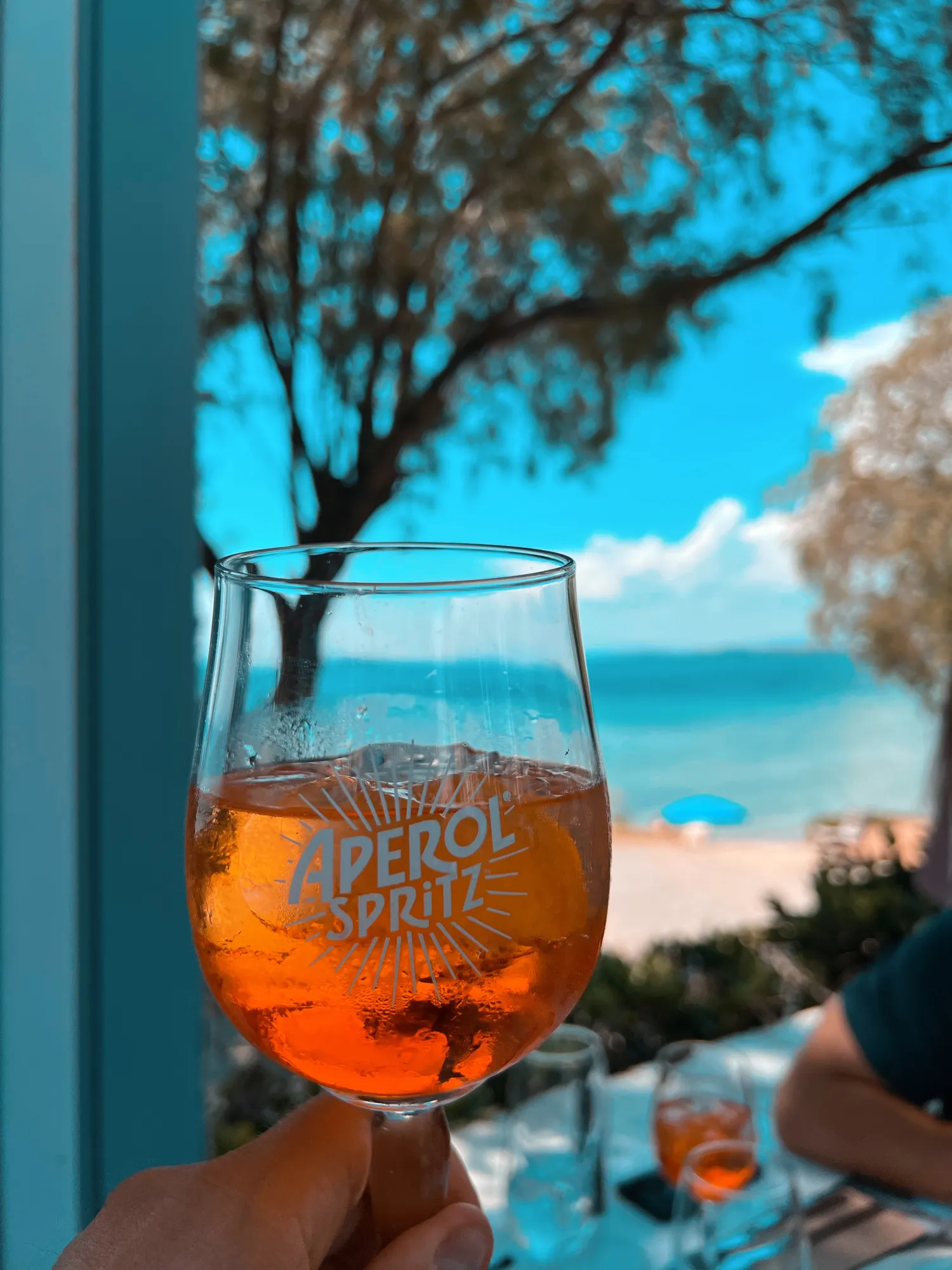 Ein Glas Aperol Spritz wird vor einem sonnigen Strand mit blauem Himmel gehalten